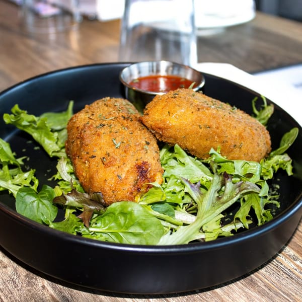 Two golden-brown arancini on a bed of mixed greens with a side of red dipping sauce.