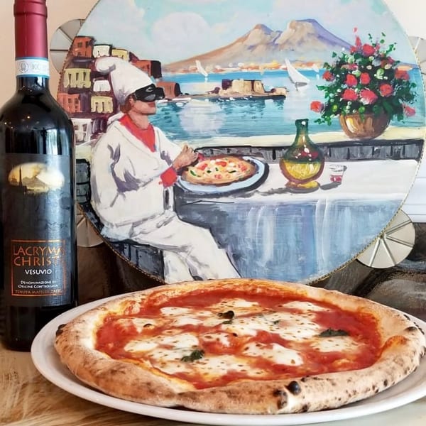 A Neapolitan pizza and a bottle of red wine in front of a painting of the Bay of Naples.